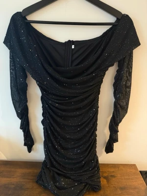 Svart glittrig offshoulder klänning - Snygg svart kortklänning med glittriga detaljer och offshoulder-design. Klänningen har långa ärmar och rynkade partier längs hela kroppen för en smickrande passform. Perfekt för dig som vill sticka ut med en trendig look.