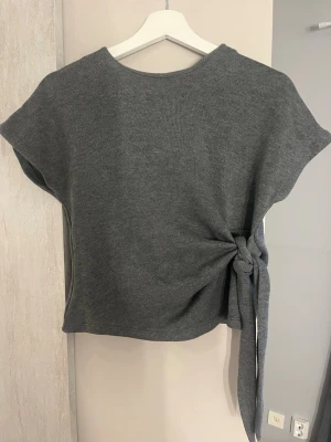 Grå stickad topp med knyt - Snygg grå stickad topp med korta ärmar och knyt i sidan som ger en cool detalj. Perfekt till jeans eller kjol för en avslappnad men trendig look. Mjuk och skön kvalitet.