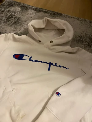 Vit hoodie från Champion - Säljer en vit hoodie från Champion med stor blå logga och röd detalj på bröstet samt ett litet Champion-märke på ärmen. Hoodien har huva med dragsko och känguruficka framtill. Perfekt för en avslappnad och sportig stil.