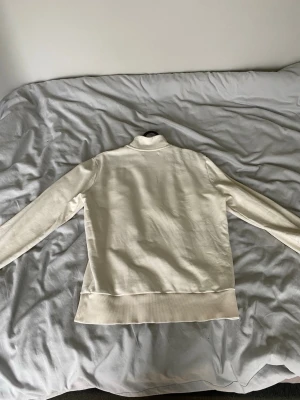 Vit halfzip-tröja från Fred Perry - Snygg vit långärmad tröja från Fred Perry med halfzip och liten broderad logga på bröstet. Tröjan har ribbade muddar och krage, samt diskreta ränder i orange och gult på ärmsluten. Perfekt för en clean och stilren look. Nypris 1300