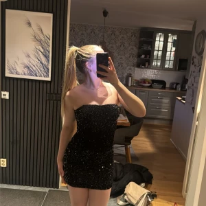 Svart glittrig axelbandslös klänning - Snygg svart axelbandslös kortklänning med glittriga paljetter över hela tyget. Klänningen har en tight passform. Stl 34 men skulle säga att den är mer 36-38. Den är däremot stretch så den passar nog från 34-38