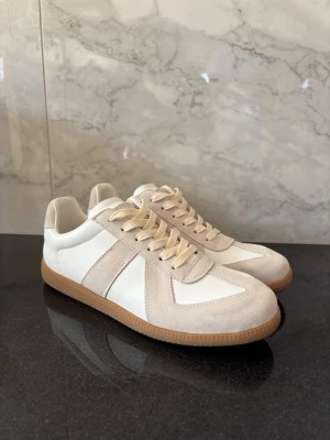 Maison Margiela gats - Snygga sneakers i vitt och beige från Maison Margiela med klassisk design. Ovandel i mjukt läder och mocka, beigea detaljer och matchande snörning. Yttersula i gummi för bra grepp. Perfekta för en stilren och trendig look.                                                 (FEJK)