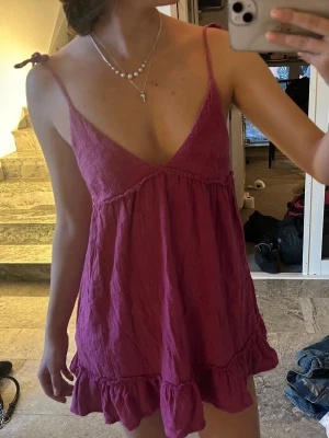 Rosa kort klänning med volang - Söt rosa kortklänning med volangkant nedtill och tunna axelband som knyts på axlarna. Djup v-ringning framtill och öppen rygg ger en avslappnad och somrig vibe. Perfekt för varma dagar och enkel att styla med smycken.