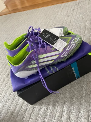 Adidas F50 lila/gröna fotbollsskor - Säljer ett par Adidas F50 fotbollsskor i en riktigt snygg lila och grön färgkombination med vita detaljer. Skorna har en extremt låg vikt, snörning och en modern design med syntetmaterial för bästa känsla på planen. Kommer med matchande skopåse och Speed innesulor. 