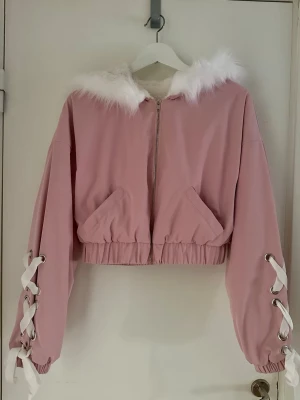 Rosa cropped vindjacka med fuskpäls - Söt rosa vindjacka med vit fuskpäls runt huvan och snörningsdetaljer med vita band längs ärmarna. Jackan är croppad, har dragkedja framtill, två snedställda fickor och resår i midjan. Perfekt för dig som gillar gullig och trendig stil.