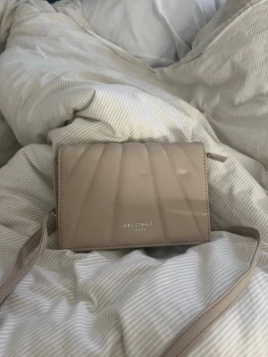 Beige axelväska från Art Street - Snygg beige axelväska från Art Street London i glansigt material med quiltade detaljer framtill. Väskan har justerbar axelrem och stilren design med guldfärgad logga på framsidan. Perfekt för dig som gillar minimalistisk och trendig look.