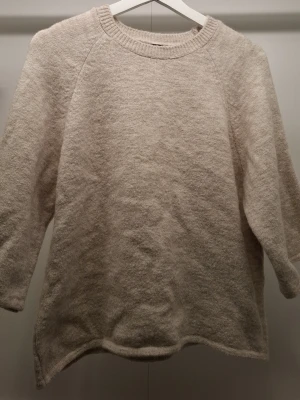 Beige oversized stickad tröja - Mysig beige stickad tröja med avslappnad passform och rund halsringning. Tröjan har långa ärmar och ribbstickade detaljer vid halsen. Perfekt för lager på lager och enkel att matcha med olika outfits.