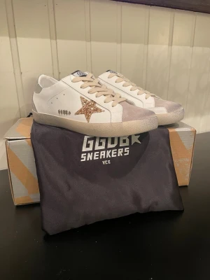 Golden Goose sneakers med glitterstjärna - Snygga vita sneakers från Golden Goose med silvriga detaljer och en glittrig stjärna på sidan. Skorna har snörning, platt sula och inslag av mocka vid tån och hälen. Perfekta för dig som vill sticka ut med en cool och trendig look.