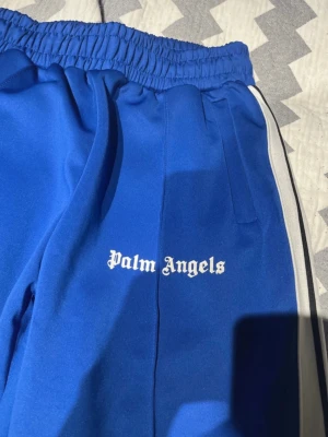 Blåa Palm Angels mjukisbyxor - Storlek M Snygga blåa mjukisbyxor från Palm Angels med vit logga på benet och vita ränder längs sidorna. Byxorna har elastisk midja och raka ben, perfekta för en chill och trendig look. Materialet känns mjukt och bekvämt mot huden.