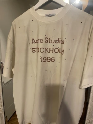 Vit Acne Studios t-shirt med strass - Säljer en vit t-shirt från Acne Studios med tryck 'STOCKHOLM 1996' framtill och små strassdetaljer över hela tröjan. Klassisk passform och rund halsringning. Perfekt för dig som gillar stilrena plagg med en twist.