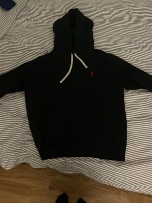 Svart hoodie från Polo Ralph Lauren - Säljer en svart hoodie från Polo Ralph Lauren med vit dragsko och den klassiska röda loggan broderad på bröstet. Hoodien har en rymlig huva och är gjord i mjukt material som passar perfekt till chill dagar. Enkel och stilren design som funkar till det mesta.