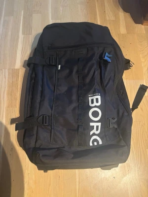 Svart ryggsäck från Björn Borg - Rymlig svart ryggsäck från Björn Borg med stor vit logga på framsidan. Utrustad med flera dragkedjefack, justerbara axelband och spännen på sidorna. Tillverkad i slitstarkt tygmaterial och har en sportig, modern look.