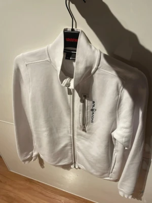 Vit zip hoodie från Sail Racing - Säljer en stilren vit hoodie från Sail Racing med dragkedja framtill och hög krage. Hoodien är Tillverkad i mjukt material som känns skönt mot huden.