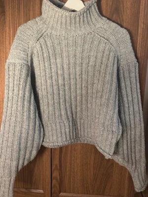 Grå ribbstickad polotröja - Säljer en grå ribbstickad polotröja med breda ärmar och croppad passform. Tröjan har hög kr hyper och är gjord i ett mjukt, tjockt garn som känns riktigt cozy. Perfekt för kyliga dagar och enkel att använda till jeans eller kjol.