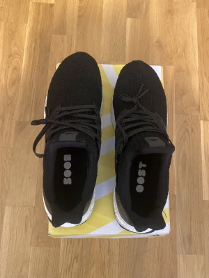 Svarta Adidas Ultra Boost sneakers - Svarta Adidas Ultra Boost sneakers med stickad ovandel och klassiska tre ränder på sidan. Skorna har en vit Boost-mellansula för extra komfort och svart yttersula. Snygg och sportig design med snörning och låg profil.