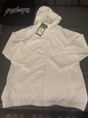 Vit träningsjacka från Under Armour - Snygg vit träningsjacka från Under Armour med huva och hel dragkedja. Jackan har diskret logga på bröstet och stor logotyp på ryggen. Tillverkad i lätt och funktionellt material, perfekt för träning utomhus. Fickor på sidorna och sportig passform. Köpt i Tyskland.