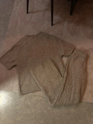 Beige stickat loungewear-set - Mysigt loungewear-set i beige stickat material med kortärmad tröja och matchande byxor. Setet har en avslappnad passform och mjuk struktur, perfekt för hemmakvällar. Materialet ser ut att vara en ull- eller mohairblandning för extra komfort.