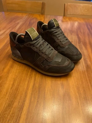 Valentino Garavani svarta sneakers - Snygga svarta sneakers från Valentino Garavani med detaljer i mocka och textil. Skorna har en klassisk låg silhuett, svarta skosnören och en beige sula med coola mönsterdetaljer. Perfekta för dig som vill ha en stilren men ändå unik look.