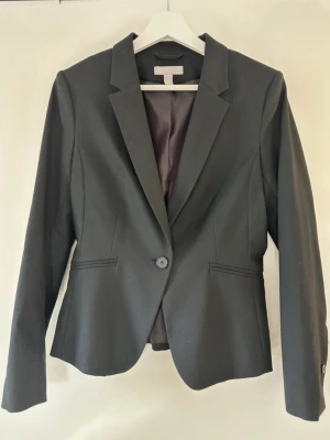 Svart klassisk kavaj med knapp - Stilren svart kavaj med en knapp framtill och två fickor. Figursydd modell med klassisk krage och helfodrad insida. Perfekt för en dressad look och passar till både byxor och kjol. Materialet är slätt och ger en snygg siluett. OBS liten i storlek sitter som en S