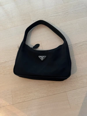 Svart axelremsväska  - Snygg svart axelremsväska klassisk trekantig logga framtill. Väskan har en enkel, stilren design med dragkedja upptill och bred rem som gör den bekväm att bära. Perfekt för dig som gillar tidlös och trendig stil.