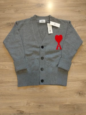 Grå cardigan från Ami med rött hjärta - Grå cardigan från Ami med djup V-ringning och tre knappar framtill. På vänster bröst sitter ett stort rött hjärta med bokstaven A. Tröjan har ribbstickade muddar och nederkant, och är tillverkad i mjukt stickat material.