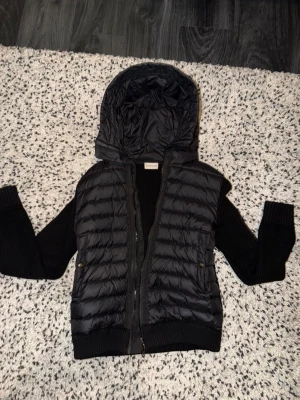 Monclear cardigan  - Snygg svart dunväst från Moncler med stickade svarta ärmar och stor huva. Västen har dragkedja framtill, två fickor med tryckknappar och Moncler-logga på ena ärmen. Materialmix av dun och stickat ger en cool och modern look.