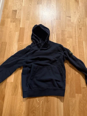 Mörkblå hoodie Stone Island M - Säljer en mörkblå hoodie från Stone Island med klassisk logga på vänster ärm. Hoodien har en stor känguruficka framtill och ribbade muddar. Perfekt för dig som gillar streetwear och vill ha något stilrent med ikonisk detalj. Storlek M