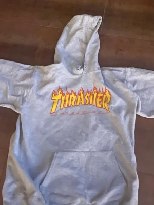 Grå Thrasher hoodie med flames - Säljer en grå hoodie från Thrasher med det klassiska flames-trycket i gult och rött på bröstet. Hoodien har huva med dragsko och en stor magficka framtill. Perfekt för dig som gillar streetwear och skatekultur.