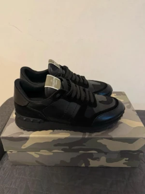Svarta Valentino Garavani sneakers - Snygga svarta sneakers från Valentino Garavani med kamouflagemönster och detaljer i läder och mocka. Skorna har svarta skosnören, robust sula och logga på plösen. Perfekta för dig som vill ha en stilren och exklusiv look.