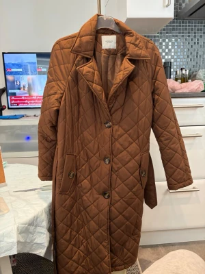 Brun quiltad kappa från Arket - Stilren brun kappa från Arket i quiltat material med rutmönster. Lång modell med klassisk krage, knäppning framtill och två fickor med lock. Perfekt för höst och vinter när du vill ha en trendig och varm look.