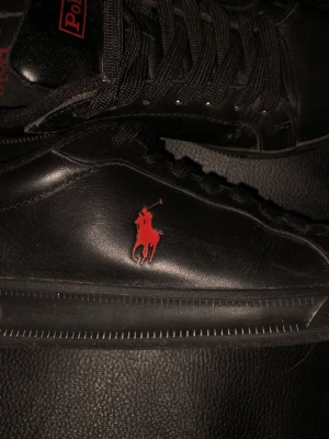 Svarta Polo Ralph Lauren sneakers - Svarta sneakers i läder från Polo Ralph Lauren med svart sula, svarta skosnören och röd Polo-logga på sidan. Klassisk låg modell med diskret design och röd Polo-märkning på plösen. Perfekta för en stilren och clean look.