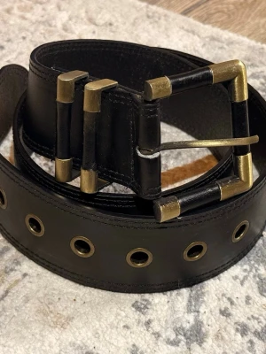 Svart bälte med metallspänne - Snyggt svart bälte i skinn med bred design och stora metallöljetter längs hela bandet. Spännet är vintage och har dubbla metallhållare för extra detalj. Perfekt accessoar för att lyfta din outfit.