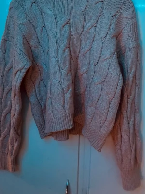 Ljusrosa kabelstickad tröja - Mysig rosa kabelstickad tröja med flätat mönster och långa ärmar. Tröjan har ribbstickade muddar vid ärmslut och nederkant, och är perfekt för lager-på-lager-stil. Materialet känns mjukt och värmande, perfekt för kyliga dagar.