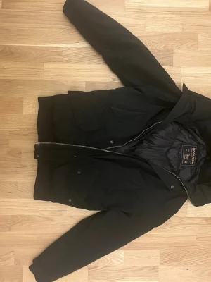 Svart bomberjacka från Woolrich - Snygg svart bomberjacka från Woolrich med klassisk passform och ribbade muddar. Jackan har dragkedja framtill, två stora fickor med lock och tryckknappar samt en mjuk, quiltad insida. Perfekt för dig som vill ha en stilren och tidlös look.