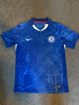 Chelsea FC matchtröja Nike blå - Chelsea FC matchtröja i blått med vita och röda detaljer. Tröjan har klubbmärke på bröstet, Nike-logga och mönstrad design. Tillverkad i lätt och ventilerande material, perfekt för fotbollsträning eller supporterliv.