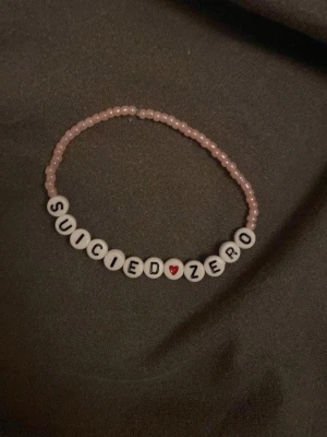 Pärlarmband med text Suicide Zero - Sött armband med små rosa pärlor och vita bokstavspärlor som bildar texten 'SUICIDE ZERO' med ett rött hjärta i mitten. Perfekt accessoar för att visa stöd och sprida budskap. Elastiskt band för enkel användning.