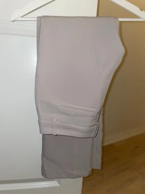 Beige kostymbyxor med pressveck - Snygga beige kostymbyxor med klassiskt pressveck och bakficka med lock. Byxorna har en rak passform och är perfekta för en stilren look. Materialet känns mjukt och följsamt, vilket ger en bekväm känsla hela dagen.