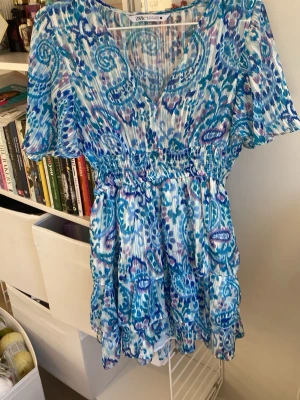 Blåmönstrad kort klänning från Zara - Superfin kort klänning från Zara med blått och lila abstrakt mönster. Klänningen har v-ringning, korta ärmar och smock i midjan för en snygg passform. Volangdetaljer nertill ger en extra touch. Perfekt för sommarens alla tillfällen!