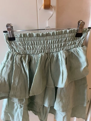 Ljusgrön volangkjol med resårmidja - Superfin ljusgrön kjol med volanger och bred smockad resår i midjan. Kjolen har en luftig och flowy känsla, perfekt för varmare dagar. Materialet känns mjukt och lätt mot huden. Om man vill ha fler bilder på plagget (på eller andra vinklar) är det bara att säga till så fixar jag det
