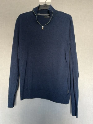 Mörkblå stickad tröja Jack & Jones - Mörkblå stickad tröja från Jack & Jones med halv dragkedja vid kragen. Tröjan har långa ärmar, ribbade muddar och nederkant. Perfekt för lager-på-lager och enkel att matcha med jeans eller chinos.