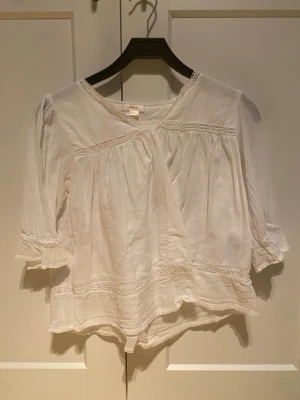 Vit romantisk blus med volang - Luftig vit blus i romantisk stil med volangdetaljer och rynkade partier. Blusen har trekvartsärm med lätt puff och dekorativa sömmar. Tillverkad i ett tunt, vävt material som ger en bohemisk känsla. Perfekt för en avslappnad men ändå söt look.