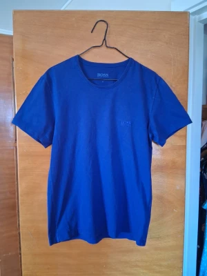 Blå t-shirt från BOSS - Säljer en stilren blå t-shirt från BOSS med diskret logga på bröstet. T-shirten har rund hals och korta ärmar, perfekt för dig som gillar klassiska och cleana plagg. 😃