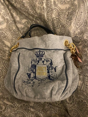 Ny Grå Juicy Couture väska - Stilren grå handväska från Juicy Couture i mjukt tyg med mörkblå detaljer och guldfärgade kedjehandtag. Väskan har broderad logga och krona framtill samt dekorativa tags. Prislappar sitter kvar, ny pris 198 dollar.