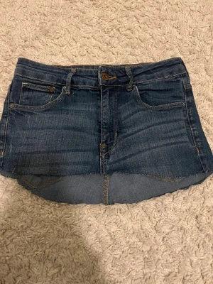 Mörkblå jeanskjol från 8Denim - Snygg mörkblå jeanskjol från 8Denim med klassisk femficksdesign och rå kant nertill. Kjolen har bälteshällor, knapp- och dragkedjestängning samt bakfickor med läderpatch. Perfekt till sommaren för en avslappnad look.