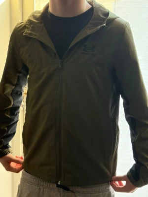 Olivgrön vindjacka från Under Armour - Snygg olivgrön vindjacka från Under Armour som är i bra skick, den har en dragkedja som går hela vägen upp till luvan . Jackan har diskret logga på bröstet och svarta detaljer längs ärmarna. Perfekt för blåsiga dagar och sportiga outfits.