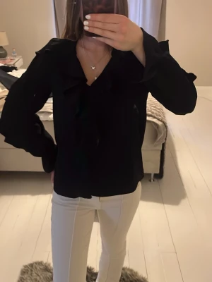 Svart blus med volangdetaljer - Stilren svart blus med långa ärmar och snygga volangdetaljer längs kragen och framtill. Blusen har en lös passform och är tillverkad i ett lätt, vävt material som ger ett elegant intryck. Perfekt att matcha med både jeans och kjol.