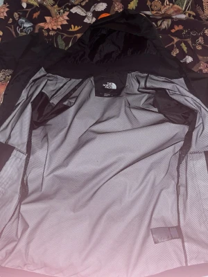 Svart vindjacka från The North Face - Snygg svart vindjacka från The North Face med huva och dragkedja framtill. Jackan har en minimalistisk design med diskret logga på bröstet och är tillverkad i lätt syntetmaterial. Perfekt för blåsiga dagar och enkel att packa ner i väskan. Passar M, L. Nypris runt 2000. Priser kan diskuteras och öppen för byten