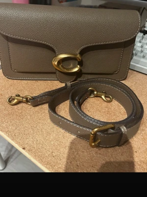 Coach brun axelremsväska i läder - Snygg brun handväska från Coach i grained läder med avtagbar och justerbar axelrem. Väskan har ett stort guldfärgat C-spänne framtill och stilrena detaljer. Perfekt för dig som gillar klassisk och tidlös design med en modern touch.