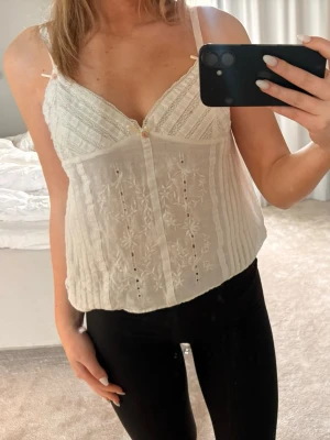 Vit spetsig linnetopp med broderi - Superfin vit linnetopp med tunna axelband, spetsdetaljer och broderade blommor framtill. Toppen har v-ringning och är lätt och luftig, perfekt för varma dagar. Söt och feminin stil som passar till både jeans och kjol.