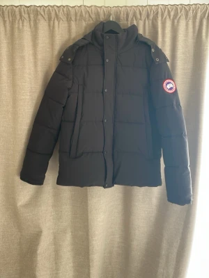 Svart pufferjacka från Canada Goose - Säljer en svart pufferjacka från Canada Goose med klassisk logotyp på ärmen. Jackan har hög krage, avtagbar huva och stora fickor framtill. Perfekt för kalla dagar med sin tjocka vaddering och stilrena design.